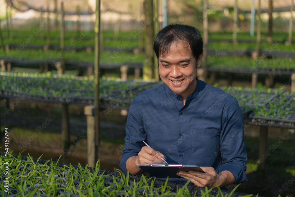 Young man farmer working at his orchid farm. Young man notes the information on the notepad to track the growth of orchid. Portrait asian small business owner of orchid gardening farm. Young man farmer working at his orchid farm. Young man notes the information on the notepad to track the growth of orchid. Portrait asian small business owner of orchid gardening farm.