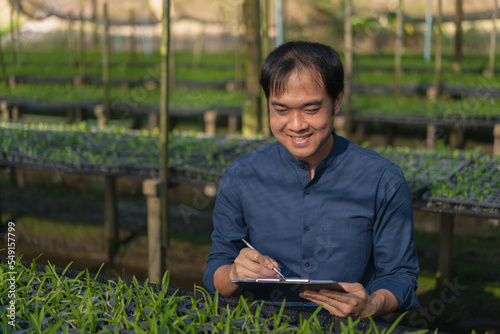 Young man farmer working at his orchid farm. Young man notes the information on the notepad to track the growth of orchid. Portrait asian small business owner of orchid gardening farm. 