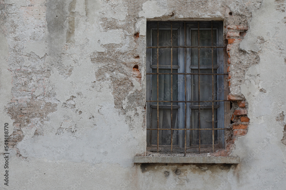Old and cracked house wall with window, frame closed and with lattice, damaged bars, wall with bricks