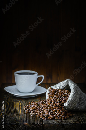 A bag of coarse canvas of coffee beans is lying on its side. Coffee beans scattered on the wooden tabletop. Coffee in white coffee cup in the background. Background with dark wood texture.