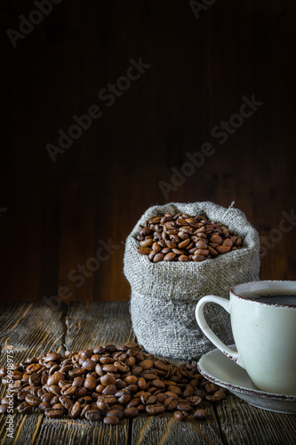 A bag of coarse canvas of coffee beans on wooden table top. Coffee beans scattered around on the wooden tabletop. Coffee in coffee cup in the foreground. Background with dark wood texture.