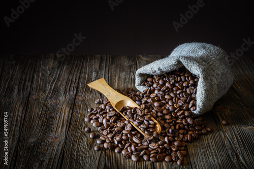 A bag of coarse canvas of coffee beans is lying on its side. Coffee beans scattered on the wooden tabletop and Coffee beans on a wooden spoon. Background with dark wood texture.