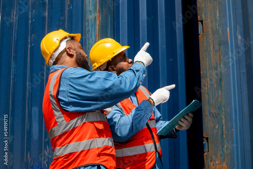 Two attractive black workers in safety helmets and uniforms work at the container port terminal. Two male engineers are checking the products in the cargo container before being exported to the port.