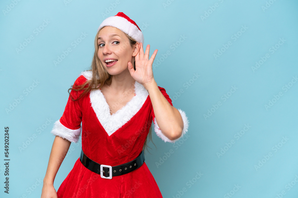 Young blonde woman dressed as mama claus isolated on blue background listening to something by putting hand on the ear Young blonde woman dressed as mama claus isolated on blue background listening to something by putting hand on the ear