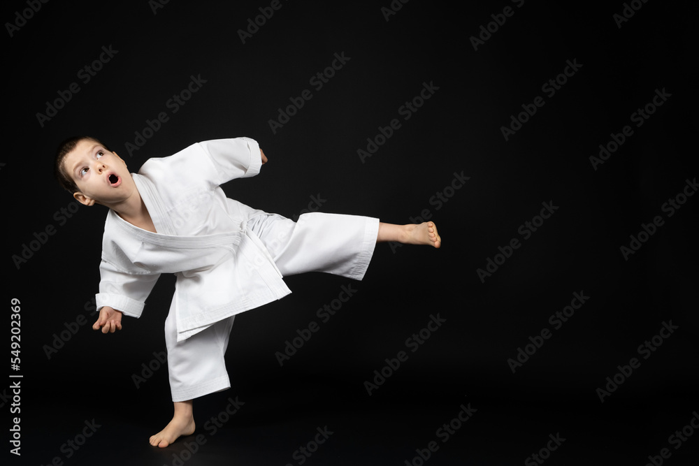 A little boy in a kimono practices karate on a black background, kicking forward. A little boy in a kimono practices karate on a black background, kicking forward.