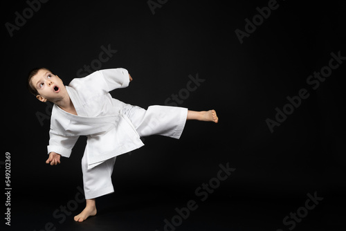 A little boy in a kimono practices karate on a black background, kicking forward.