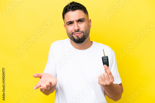 Young Arab man holding car keys isolated on yellow background making doubts gesture while lifting the shoulders