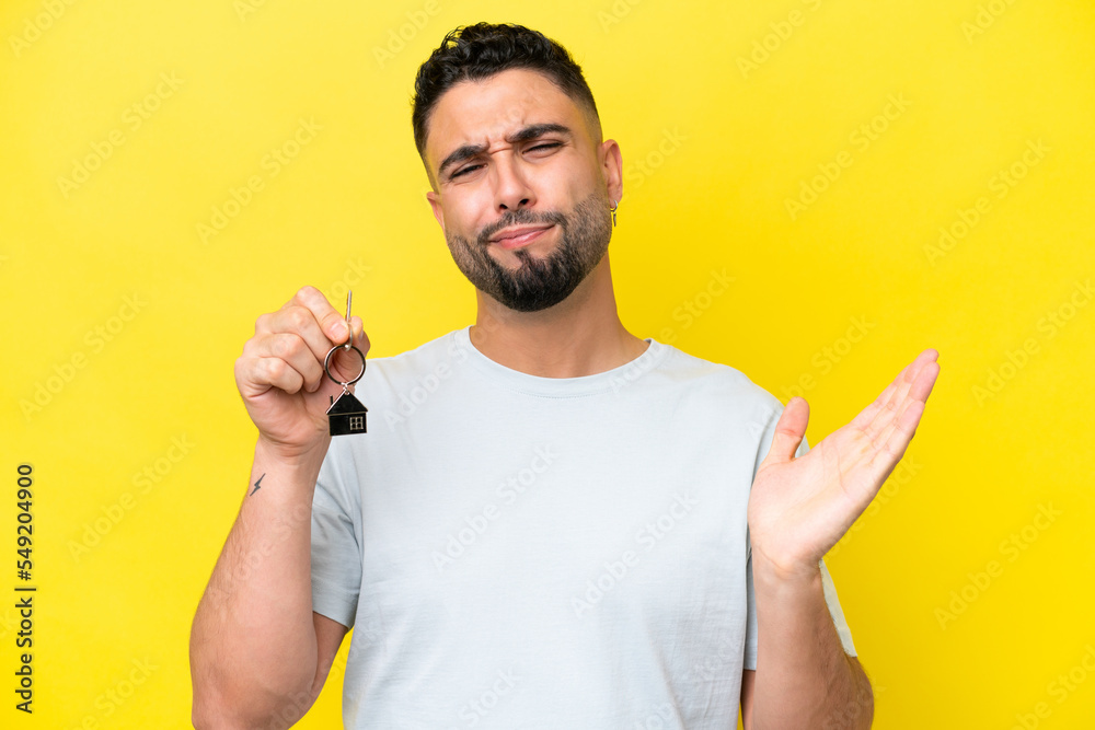 Young Arab man holding home keys isolated on yellow background making doubts gesture while lifting the shoulders Young Arab man holding home keys isolated on yellow background making doubts gesture while lifting the shoulders