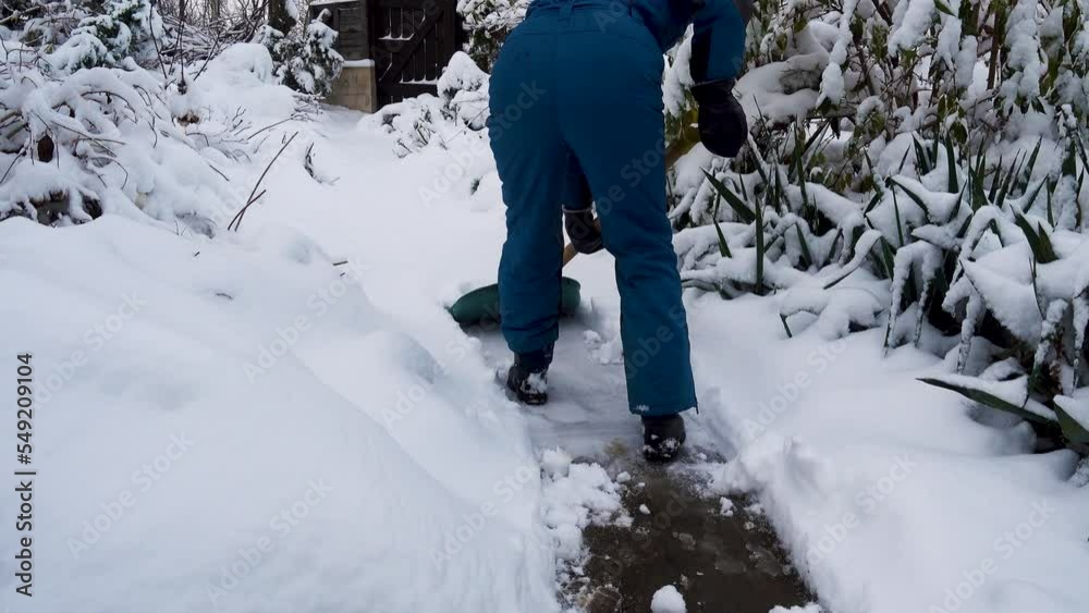 Happy young woman cleans snow in the garden near the house. Portrait of a girl with a snow shovel in her hands. Happy girl removes snow near the house in the garden. The concept of happiness, joy.