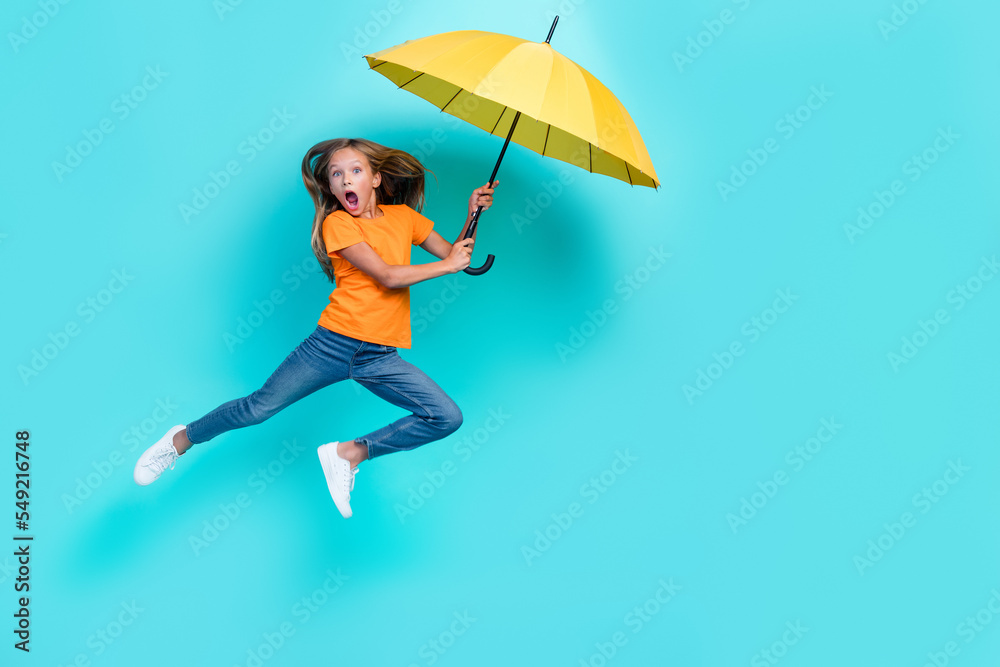 Full length photo of young charming crazy schoolkid girl jumping air hold umbrella open mouth flying air dreamy windy weather isolated on cyan color background