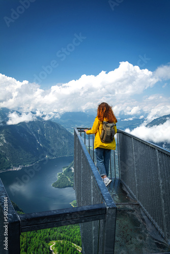 Redhead girl in yellow jacket is looking at  Hallstatter lake, Hallstatt and surrounding mountains from 5 Fingers view point in Austria. Dachstein mountains.