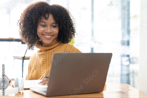 Photo of cheerful joyful mixed-race woman in yellow shirt smiling work on laptop talk speak video call online. Smart ethnic female in earphones study distant on computer at home. Education concept.