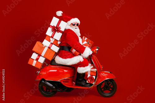 Profile side view of his he nice funny cheery white-haired St Nicholas riding moped hurry up rush delivering bringing pile stack giftboxes isolated bright vivid shine vibrant yellow color background