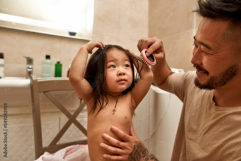 Fototapeta premium Smiling asian father combing hair of daughter