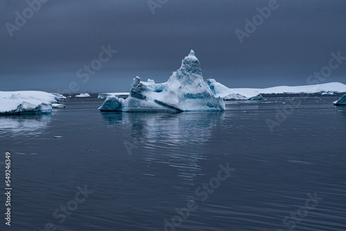 A snow-covered iceberg floating in the Antarctic Ocean, with a reflection in the water. The dark grey sky is also reflected in the water. 