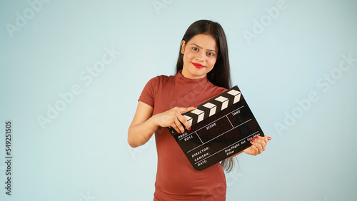 Beautiful young Asian Indian woman standing holding clapperboard, clapper board used in film making, isolated on blue color background studio portrait