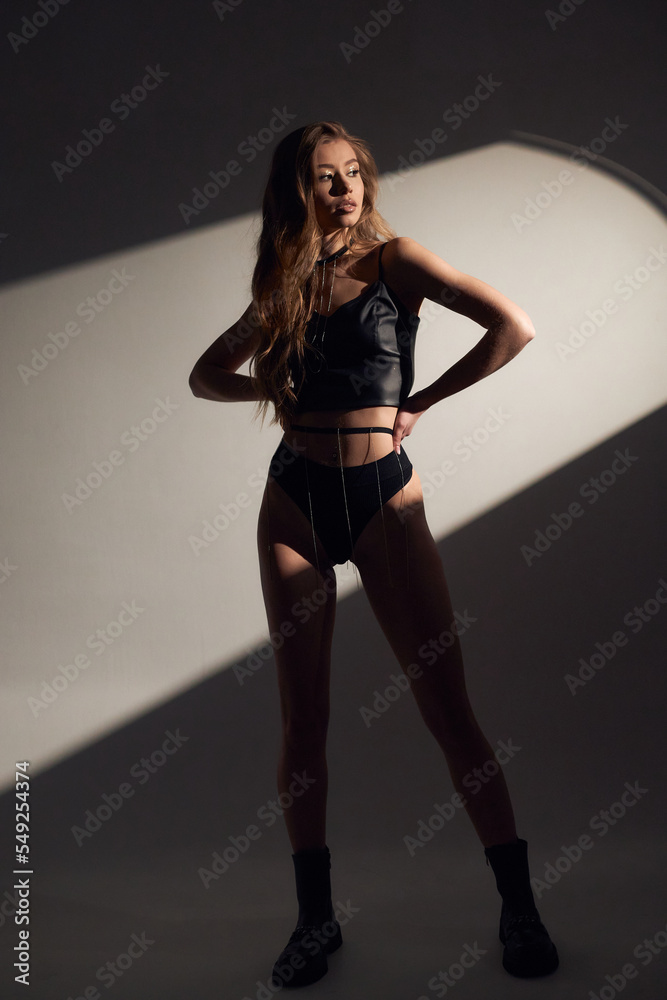 Studio portrait of young attractive woman model with brunette long curly hair, in black bodysuit dress and high heels, long legs with tan skin, slim body, stay on background with sun hard light.