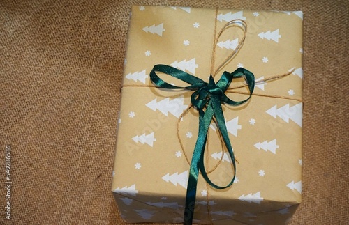 Weihnachtsgeschenk in beigegelbem Papier mit weißem Weihnachtsbaummuster und grün-goldener Schleife verpackt auf braunem Laminatboden photo