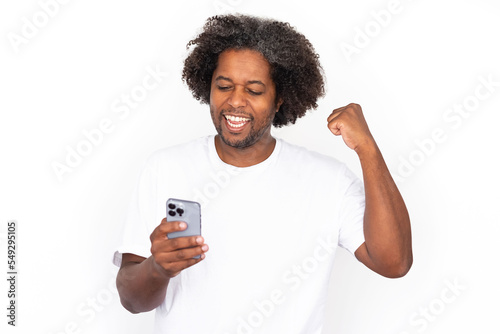 Happy African American man using phone with clenched fist. Excited mature male model with dark curly hair in white T-shirt looking at screen, winning in game. Modern technology, advertising concept