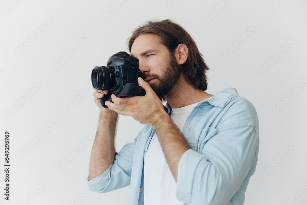 A male hipster photographer in a studio against a white background looks through the camera viewfinder and shoots shots with natural light from the window. Lifestyle work as a freelance photographer