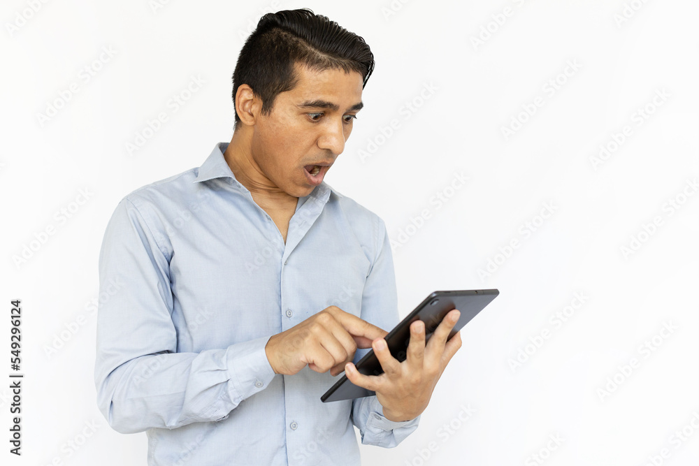 Shocked man looking at tablet. Indian man in blue shirt holding tablet and expressing surprise. Portrait, studio shot, technology, surprise concept Shocked man looking at tablet. Indian man in blue shirt holding tablet and expressing surprise. Portrait, studio shot, technology, surprise concept