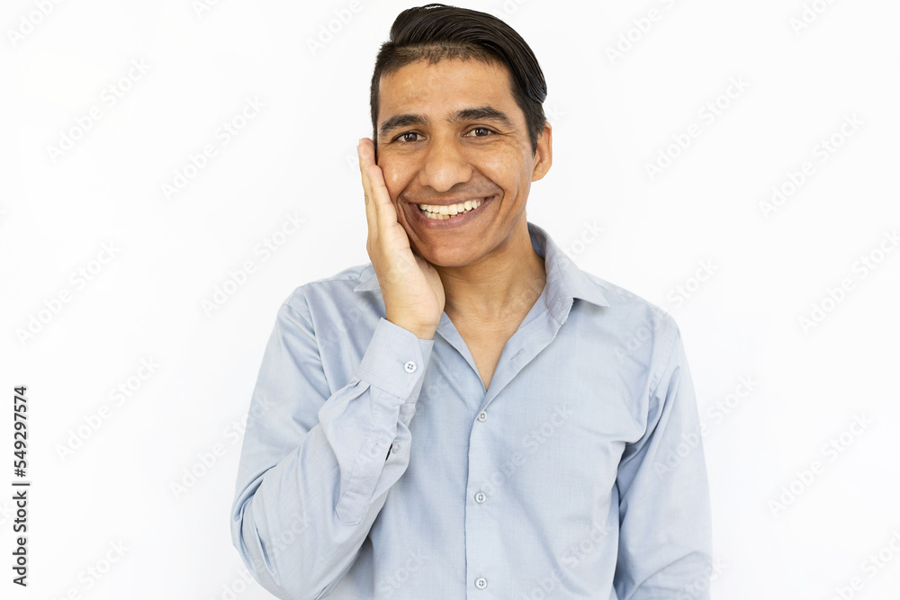 Happy man smiling at camera while standing on white background. Cheerful Indian man in shirt looking at camera with his hand on his face, receiving good news. Happiness concept