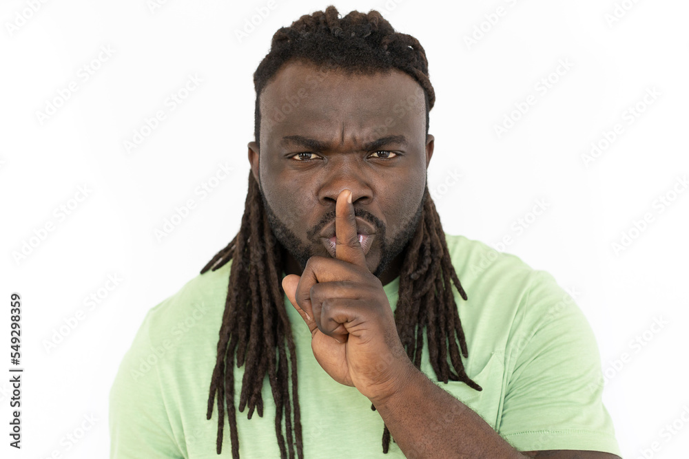 Concentrated African American man showing hush sign at camera. Front view of serious man in T-shirt looking at camera, asking to be quiet. Silence, hush, secrecy concept