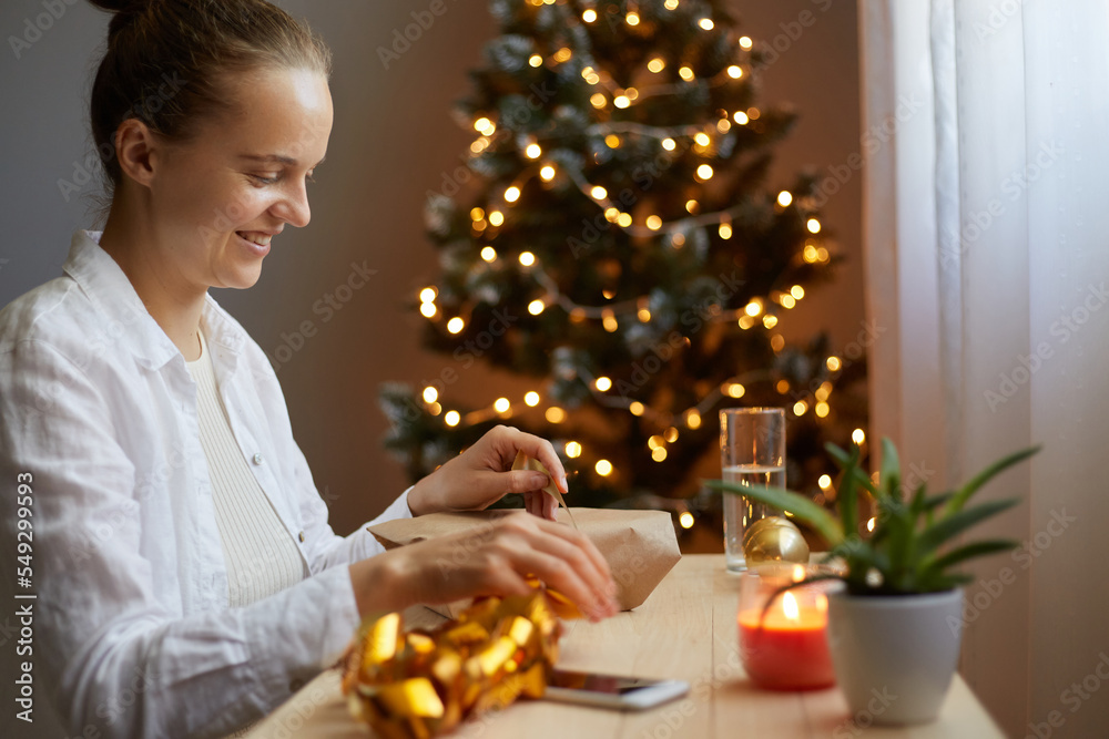 horizontal shot of smiling young adult Caucasian woman opening her present on Christmas, smiling, being happy, having good mood, packing gift fore child or husband.