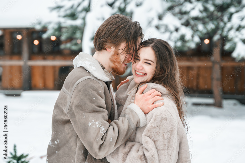 Fototapeta premium Young couple girl guy walking near barn with skates. Snowy cold winter in countryside. Skating, kissing, having fun, laughing in stylish clothes, fur coat,hat.Romantic love story,village date,weekend