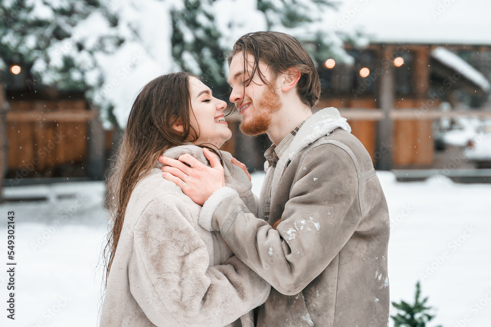 Obraz premium Young couple girl guy walking near barn with skates. Snowy cold winter in countryside. Skating, kissing, having fun, laughing in stylish clothes, fur coat,hat.Romantic love story,village date,weekend
