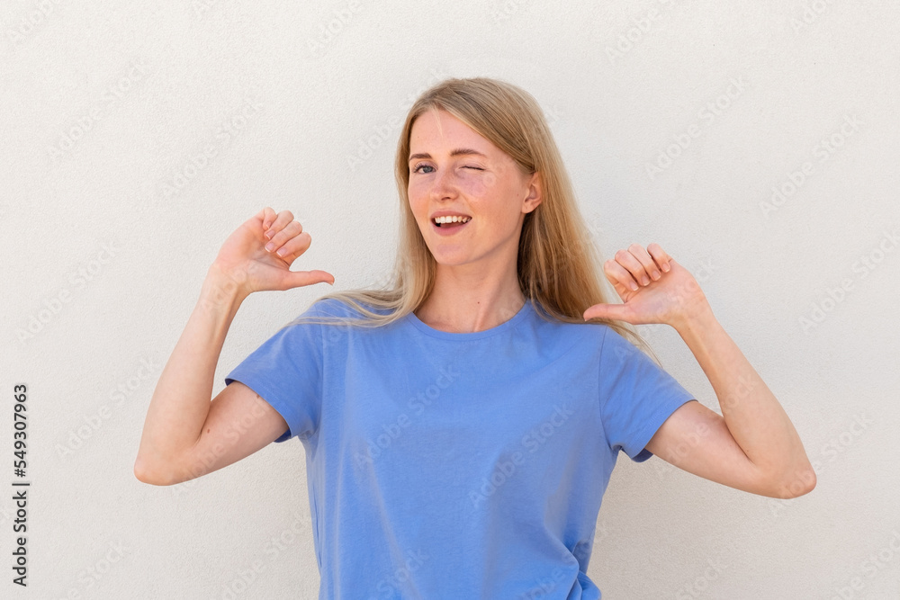 Winking young woman pointing at herself with thumbs Caucasian female model with blonde hair and blue eyes in blue T-shirt smiling and pointing at herself. Self-assurance, confidence concept Winking young woman pointing at herself with thumbs Caucasian female model with blonde hair and blue eyes in blue T-shirt smiling and pointing at herself. Self-assurance, confidence concept