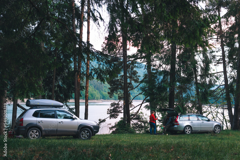 Travel and tourism by two car. Vehicle near tall coniferous trees. Tents and active recreation in the Carpathian Mountains and lake. Camping in forest.