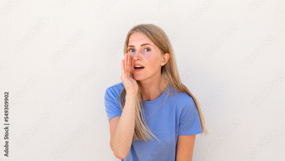 Close-up of whispering young woman holding hand near mouth. Caucasian female model with blonde hair and blue eyes in blue T-shirt telling something confidential. Secrecy, privacy concept. Close-up of whispering young woman holding hand near mouth. Caucasian female model with blonde hair and blue eyes in blue T-shirt telling something confidential. Secrecy, privacy concept.