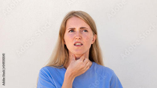 Dissatisfied young woman suffering from sore throat. Caucasian female model with blonde hair and blue eyes in blue T-shirt touching her throat feeling discomfort. Pain, soreness concept