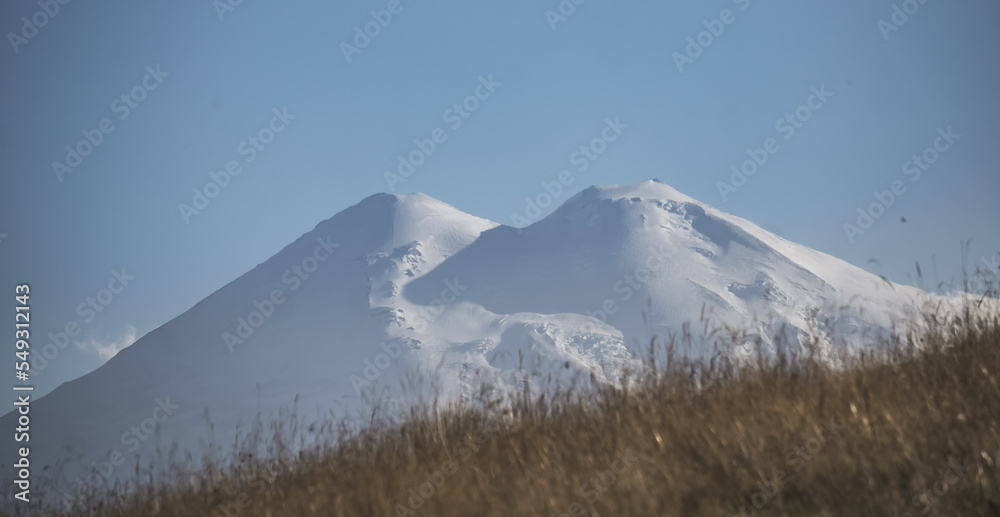 Panorama of Mount Elbrus with snow and glaciers at side evening illumination and yellowed autumn grass in the mountains, warm autumn evening in the mountains