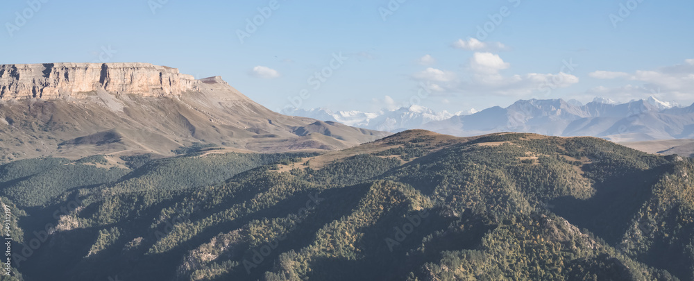 Panorama in the evening in the mountains of the Caucasus on a steep rocky slope of a table mountain against the backdrop of a mountain range with snow, hills with vegetation below