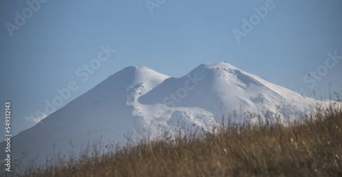 Panorama of Mount Elbrus with snow and glaciers at side evening illumination and yellowed autumn grass in the mountains  warm autumn evening in the mountains