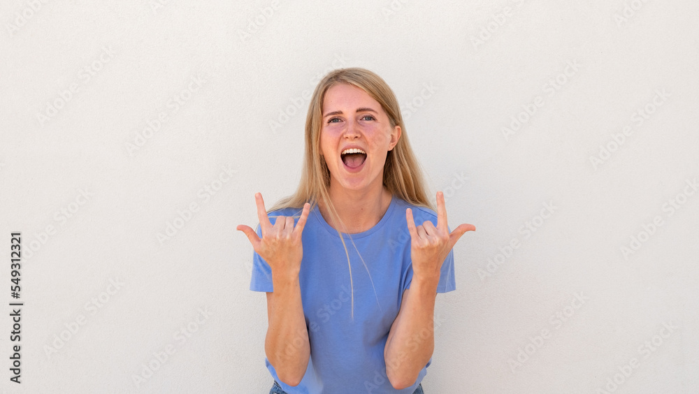 Emotive young woman with open mouth showing rock-n-roll gesture. Caucasian female model with blonde hair and blue eyes in blue T-shirt showing horns up. Lifestyle, rebellious concept Emotive young woman with open mouth showing rock-n-roll gesture. Caucasian female model with blonde hair and blue eyes in blue T-shirt showing horns up. Lifestyle, rebellious concept