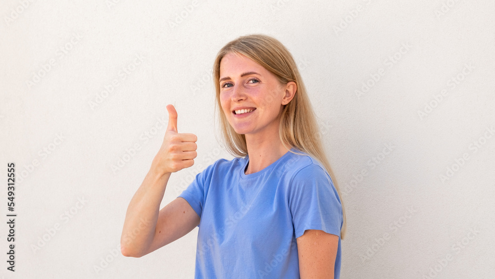 Cheerful young woman showing thumb up standing aside. Caucasian female model with blonde hair and blue eyes in blue T-shirt smiling and showing thumb up. Social media, advertisement concept Cheerful young woman showing thumb up standing aside. Caucasian female model with blonde hair and blue eyes in blue T-shirt smiling and showing thumb up. Social media, advertisement concept