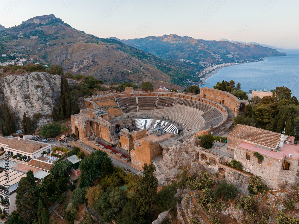 Aerial view of the Ruins of the Ancient Greek Theater in Taormina, Sicily with the double smoke tail of the Etna extending over the the Giardini-Naxos bay of the Ionian Sea in the morning sun shine.