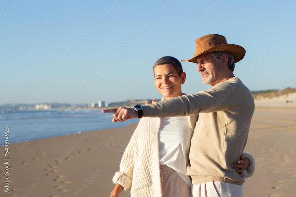 Happy senior couple standing at the beach, enjoying sea view. Grey-haired man in cowboy hat pointing at something while smiling short-haired lady holding him around waist. Leisure, love concept