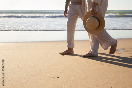 Couple enjoying vacation, walking along seashore with seascape in background. Legs closeup of lady in cardigan with straw hat and man strolling barefoot on wet sand. Romance, holiday concept