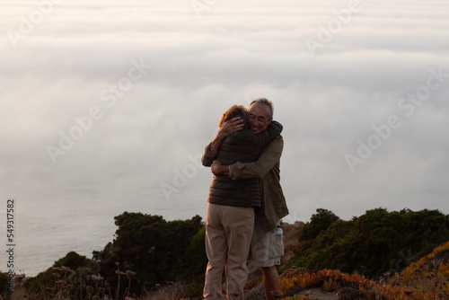 Contented elderly hikers at sunset. Man and woman in casual clothes and with ammunition standing at peak, hugging. Hobby, active lifestyle, love concept