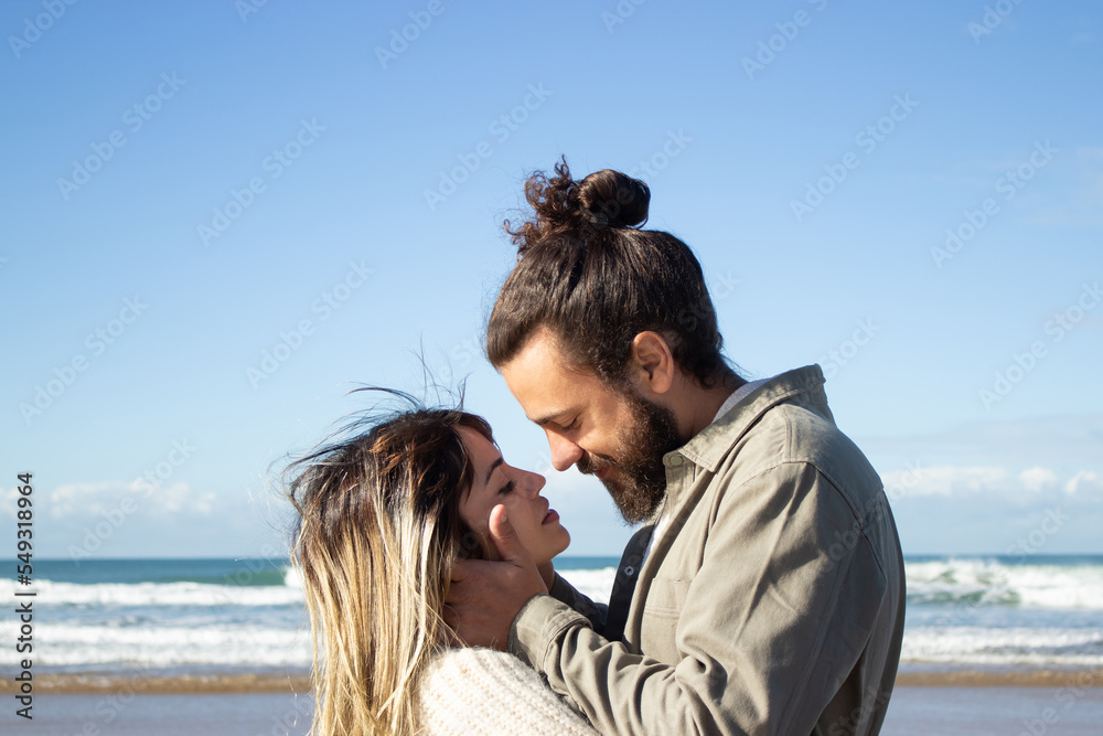 Fototapeta premium Happy couple spending time at seashore, showing affection and embracing. Loving guy with beard tenderly holding girls face, while beautiful woman with closed eyes waiting for kiss. Romance concept