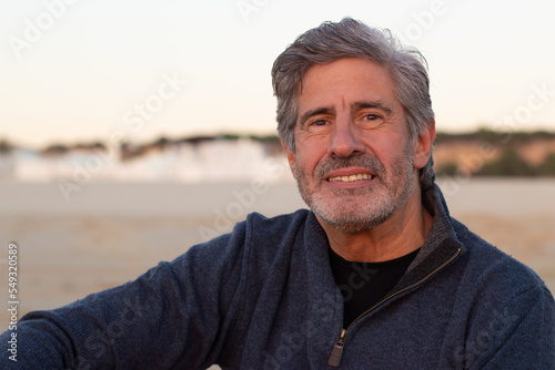 Portrait of grey-haired senior man spending time outside at seashore. Smiling middle-aged man with beard looking at camera and smiling. Medium shot. Lifestyle, retirement, leisure concept