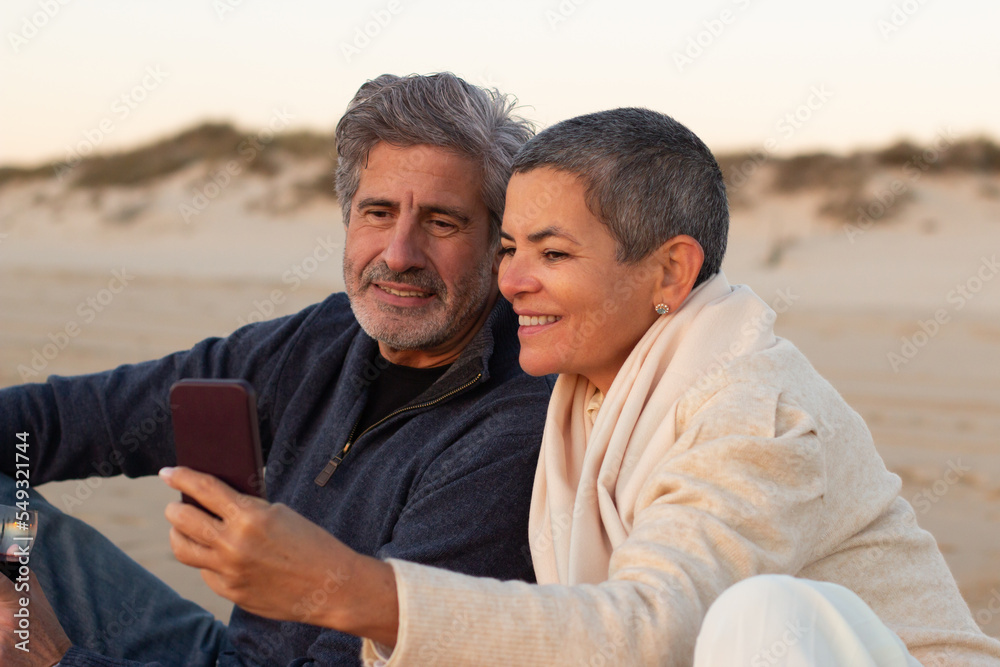 Obraz premium Senior couple spending time at the beach at sunset. Grey-haired man and smiling lady with short hair sitting on blanket and looking through photos on cellphone. Leisure, mobile communication concept