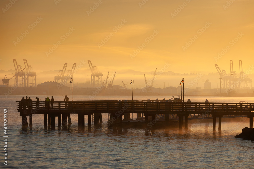 Fishing Off a Pier During During a Foggy Sunrise. With cranes for loading freighters in the background fishermen hope their luck will be good in Elliott Bay, Seattle, Washington.
