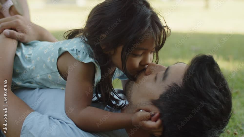 Little girl lying with father on ground and kissing him on lips. Asian young man resting on grass with little daughter in park. Fatherhood and childhood concept Stock ビデオ | Adobe Stock