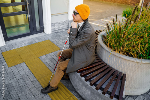 Blind man with a cane sits on a bench in the city near the tactile tiles