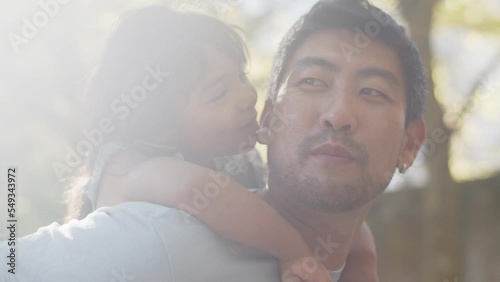 Close-up of cute little Asian girl on back of her father, embracing and kissing him. Young man smiling and talking to daughter. Fatherhood concept